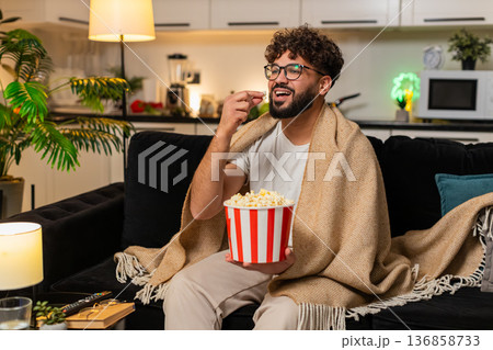 Arabian man watches movie with popcorn wrapped in blanket laughs comments on show at night indoors Arabian man watches movie with popcorn wrapped in blanket laughs comments on show at night indoors 136858733