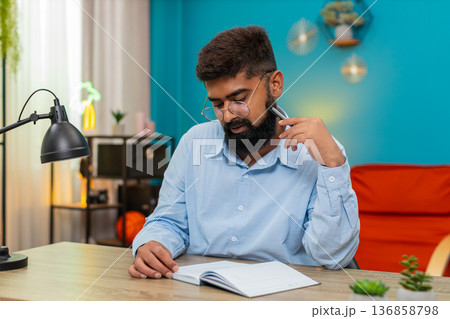 Indian man holds pen gazes notebook deeply thinking planning ideas writing strategy indoors focused 136858798