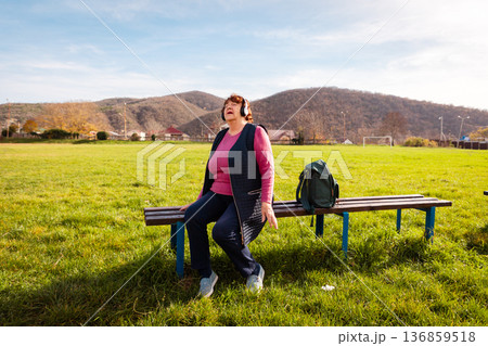 Senior Caucasian woman listening to music with headphones while relaxing in a park. She enjoys peaceful outdoor rest. Concept of happy retirement, mental wellbeing Senior Caucasian woman listening to music with headphones while relaxing in a park. She enjoys peaceful outdoor rest. Concept of happy retirement, mental wellbeing 136859518