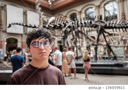 Berlin, germany, august 15, 2023. Man with glasses standing in a museum hall, observing dinosaur skeletons and other visitors. Learning and education concept 136859921