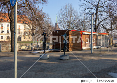 Wooden structure stands by the river with two sculptures on the sidewalk 136859936