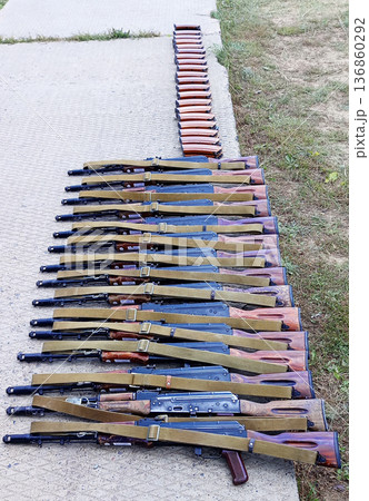 Large collection of AK-style rifles, neatly lined up on concrete next to an edge of grass, with corresponding rifle magazines stacked in a row nearby Large collection of AK-style rifles, neatly lined up on concrete next to an edge of grass, with corresponding rifle magazines stacked in a row nearby 136860292