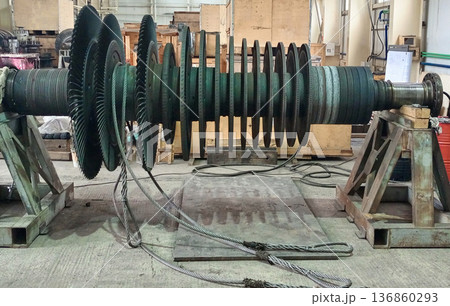 Industrial turbine rotor with multiple stages of blades rests on heavy metal supports inside a workshop, with steel cables in the foreground 136860293