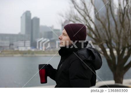Young man enjoying a warm drink from a disposable cup near the river on a chilly day in winter. Concept of a city winter activity 136860300