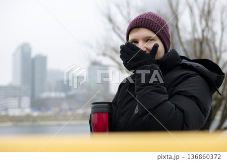 Young man enjoying a warm drink from a red travel mug near the river on a chilly day in winter. Concept of a city winter activity 136860372