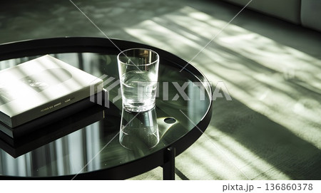 black coffee table with a glass of water and two books on it. a ray of sunlight shines through the window onto the tabletop in an elegant, dark room 136860378
