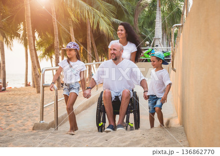 Disabled man in a wheelchair moves on a ramp to the beach with his family. 136862770