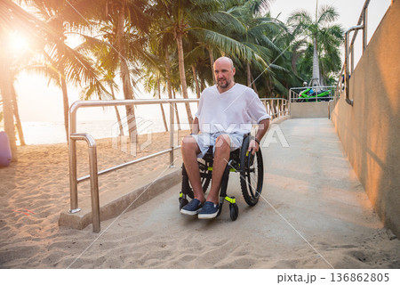 Disabled man in a wheelchair moves on a ramp to the beach. Disabled man in a wheelchair moves on a ramp to the beach. 136862805