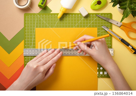 Hands draw line with pencil along ruler on yellow paper. Cutting mat, scissors, glue, and stationery. 136864014