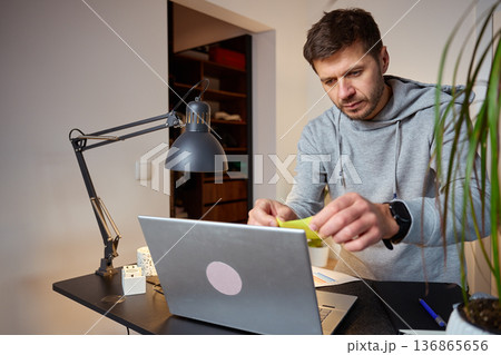 Man working at home office desk placing sticky note on laptop screen Man working at home office desk placing sticky note on laptop screen 136865656