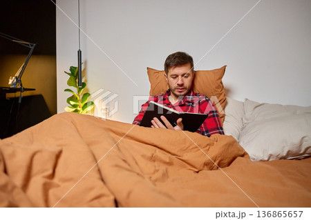 Man sitting in bed and reading book under blanket in bedroom 136865657
