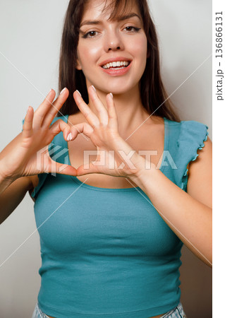 Young woman forms heart shape with fingers and smiles while standing indoors in front of a simple background Young woman forms heart shape with fingers and smiles while standing indoors in front of a simple background 136866131