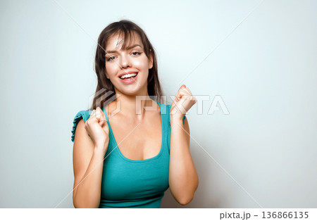 Young woman smiles and raises her hands in celebration while standing against a light background in a cheerful moment 136866135