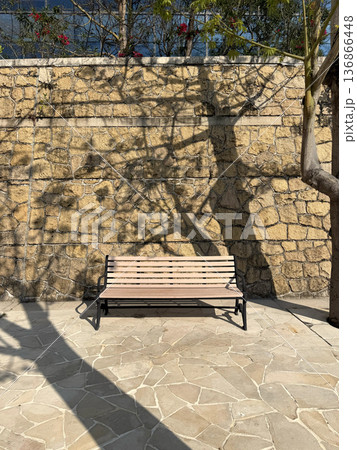 Empty wooden park bench sits against a textured stone wall with dappled sunlight and tree shadows 136866448