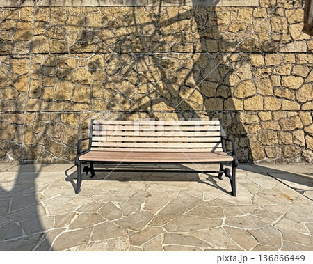 Empty wooden park bench sits against a textured stone wall with dappled sunlight and tree shadows 136866449