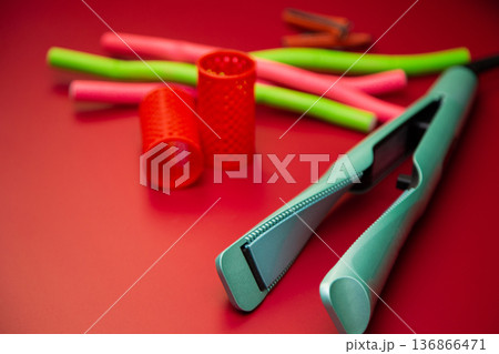 Side view of a green hair straightener with a twisted ceramic canvas on a background of curlers of different shapes and sizes on a pink background. The concept of styling, hairstyle, curls. High Side view of a green hair straightener with a twisted ceramic canvas on a background of curlers of different shapes and sizes on a pink background. The concept of styling, hairstyle, curls. High 136866471