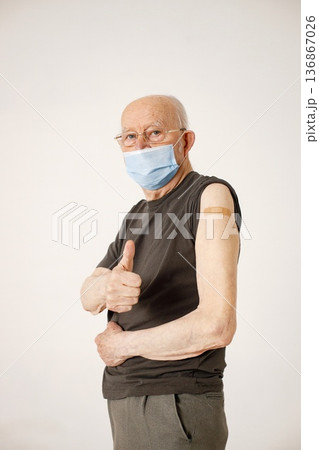 Senior gentleman on a wheelchair isolated on a white background. Man wearing khaki t-shirt and face mask. Man has a stick band-aid after vaccination and holding a thumb up. Senior gentleman on a wheelchair isolated on a white background. Man wearing khaki t-shirt and face mask. Man has a stick band-aid after vaccination and holding a thumb up. 136867026