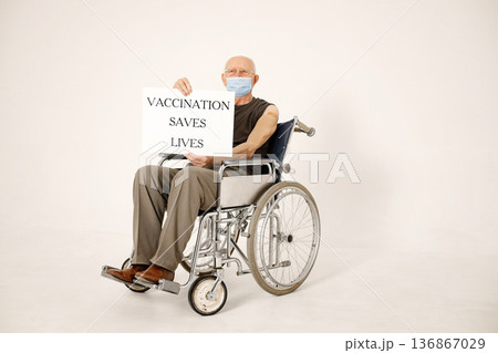 Senior gentleman on a wheelchair isolated on a white background. Man wearing khaki t-shirt and face mask. Man has a stick band-aid after vaccination and holding a poster vaccination saves lives. Senior gentleman on a wheelchair isolated on a white background. Man wearing khaki t-shirt and face mask. Man has a stick band-aid after vaccination and holding a poster vaccination saves lives. 136867029