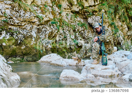 Camouflaged Sniper Soldier Resting With Tactical Rifle Beside Rocky Stream in Forest Terrain 136867098
