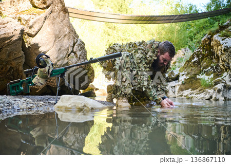 Camouflaged Sniper In Ghillie Suit By River With Tactical Sniper Rifle During Reconnaissance Camouflaged Sniper In Ghillie Suit By River With Tactical Sniper Rifle During Reconnaissance 136867110