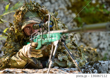 Soldier Sniper in Ghillie Camouflage Aiming a Precision Rifle From Prone Position in Woodland 136867112