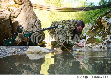 Soldier In Ghillie Suit Kneeling At Stream Drinking Water With Sniper Rifle Under Bridge Soldier In Ghillie Suit Kneeling At Stream Drinking Water With Sniper Rifle Under Bridge 136867113