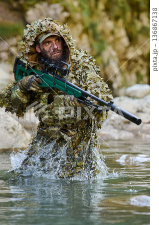 Sniper Soldier in Ghillie Suit Wading Through River With Rifle in Tactical Camouflage Sniper Soldier in Ghillie Suit Wading Through River With Rifle in Tactical Camouflage 136867138