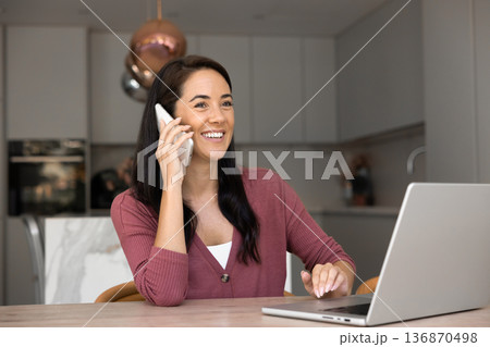 Smiling woman talking on phone seated at table with laptop 136870498