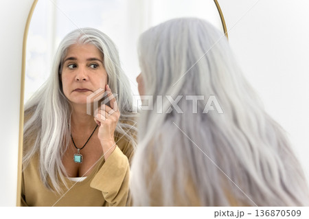 Serious middle-aged grey-haired woman looking in mirror, touch her cheekbones Serious middle-aged grey-haired woman looking in mirror, touch her cheekbones 136870509