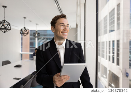 Businessman holding tablet, looks out window with positive, inspired expression Businessman holding tablet, looks out window with positive, inspired expression 136870542