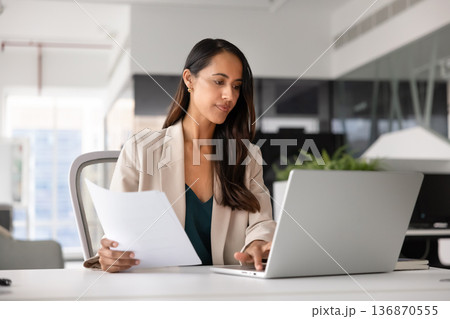 Focused businesswoman doing paperwork and working on laptop in office 136870555