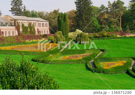 Green manicured lawn in the park, along which paths stretch, shrouded on both sides by cultivated shrubs and flowers, and extend to the house, behind which there are forest trees Green manicured lawn in the park, along which paths stretch, shrouded on both sides by cultivated shrubs and flowers, and extend to the house, behind which there are forest trees 136871280