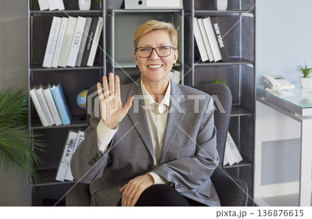 Smiling business professional waving hello during video call in modern office workspace Smiling business professional waving hello during video call in modern office workspace 136876615
