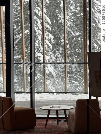 Cozy lounge seating by large window overlooking snowy pine forest and falling snow outside 136878616
