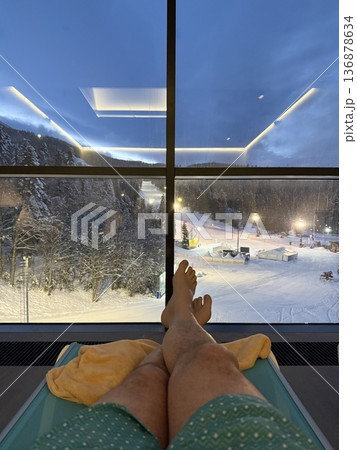 Relaxing evening view from spa lounger overlooking snowy ski resort and lit slopes through a large window 136878634
