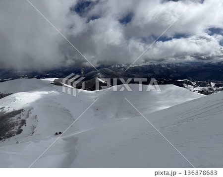 Snow-covered mountain ridge under dramatic clouds with distant valley and solitary cabin 136878683