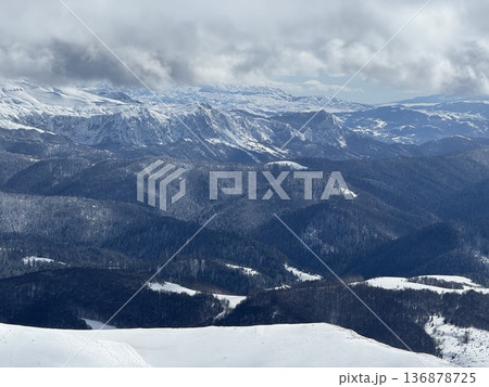 Snowy mountain range with forested ridges and valleys under a dramatic cloudy winter sky 136878725