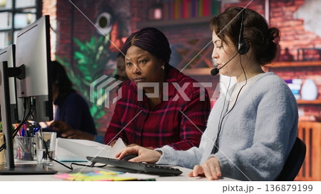 Call center team leader going over paperwork files next to colleague at PC desk. Customer support manager explaining company documentation to intern accommodating with job, camera A 136879199