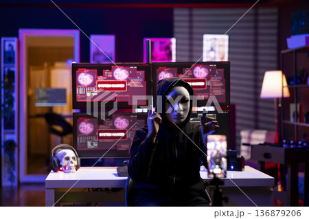 Masked cybercriminal doing espionage campaign from secret base of operations. Rogue software developer wearing anonymous mask using smartphone to record demands from secret underground lair Masked cybercriminal doing espionage campaign from secret base of operations. Rogue software developer wearing anonymous mask using smartphone to record demands from secret underground lair 136879206