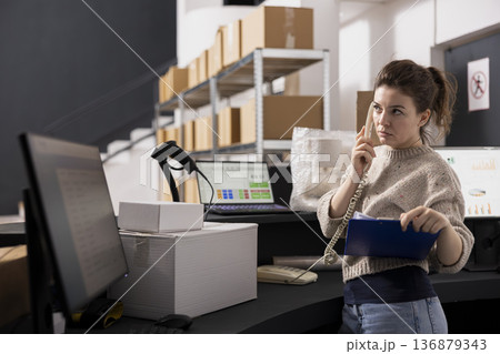 Female staff member answers a landline phone at the small scale warehouse, coordinating shipments, packaging and retail orders while managing goods, parcels and small delivery preparation. Female staff member answers a landline phone at the small scale warehouse, coordinating shipments, packaging and retail orders while managing goods, parcels and small delivery preparation. 136879343