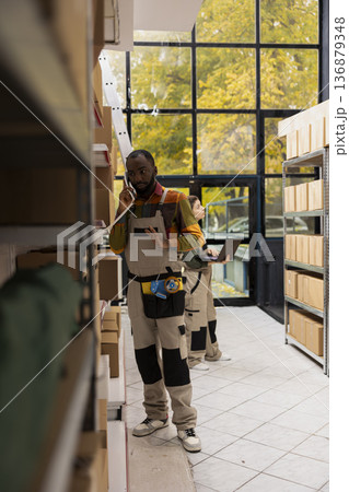 Black employee answering a call in small scale warehouse depot, confirming online orders and handling shipment service operations. Small business delivery preparation management. 136879348
