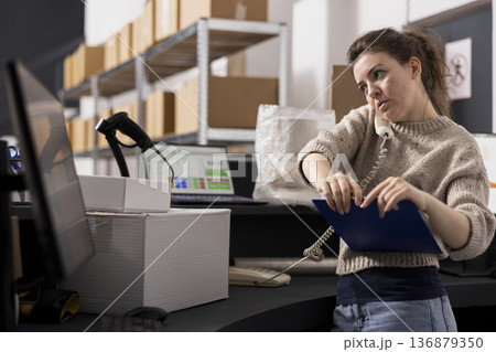 Woman worker in coveralls takes a phone call reviewing order lists and handling processing tasks for shipment service, storage facility operations and e-commerce express delivery. 136879350