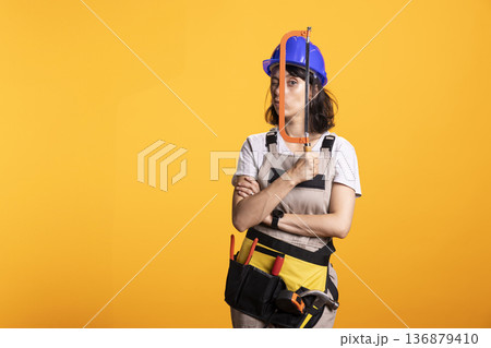 Female renovation worker poses with hacksaw tool, standing with arms crossed and playful facial expression. Confident repairwoman wearing helmet holds hacksaw in front of eye for humorous expression. 136879410