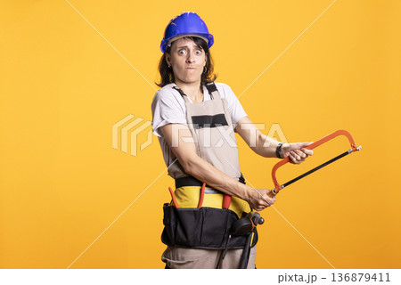 Serious white woman in construction gear uses handsaw, portraying home improvement or labor effort. Female contractor stands in safety helmet and tool apron against isolated yellow background. Serious white woman in construction gear uses handsaw, portraying home improvement or labor effort. Female contractor stands in safety helmet and tool apron against isolated yellow background. 136879411