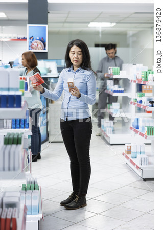 Middle aged woman examines sunscreen and heart supplements in medical retail store. Customer compares pharmaceutical products, reflecting mindful healthcare purchasing and wellness planning. 136879420