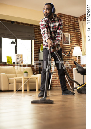 African American man enjoying music through headphones while vacuuming living room floor, completing weekend chores. Relaxed black male individual tidying up apartment with vacuum on sunny afternoon. African American man enjoying music through headphones while vacuuming living room floor, completing weekend chores. Relaxed black male individual tidying up apartment with vacuum on sunny afternoon. 136879433
