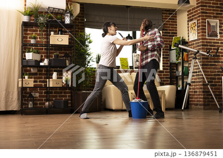 Carefree diverse couple bond while tidying their home, playfully using TV remote and mop stick as microphones. African american and caucasian spouses singing together and having fun with housework. Carefree diverse couple bond while tidying their home, playfully using TV remote and mop stick as microphones. African american and caucasian spouses singing together and having fun with housework. 136879451