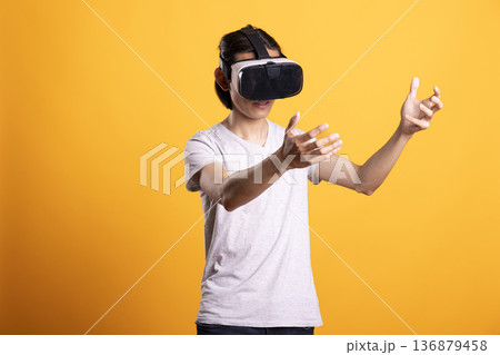 Young asian man experiencing augmented reality environment with VR glasses. Male tech enthusiast reaching forward, enjoying interactive entertainment and futuristic digital simulation. 136879458