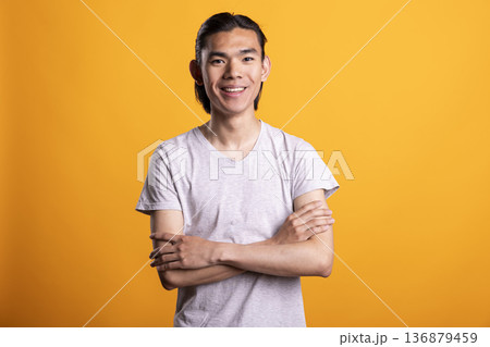 Smiling young Asian man stands confidently with folded arms, showing happiness and fulfillment. Proud and satisfied male model poses, radiating positive attitude and cheerful self assurance. Smiling young Asian man stands confidently with folded arms, showing happiness and fulfillment. Proud and satisfied male model poses, radiating positive attitude and cheerful self assurance. 136879459