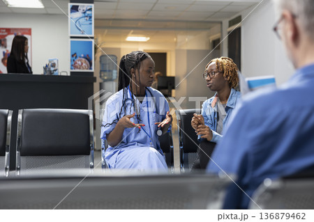Black nurse in uniform sits on bench beside patient, discussing medical tests and procedures in hospital lobby. Healthcare worker gives friendly advice to young woman about maintaining good health. Black nurse in uniform sits on bench beside patient, discussing medical tests and procedures in hospital lobby. Healthcare worker gives friendly advice to young woman about maintaining good health. 136879462